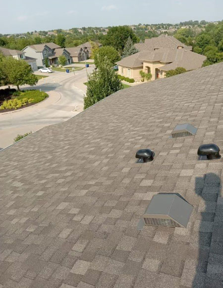 Residential roofing with asphalt shingles, featuring ventilation pipes, overlooking a neighborhood with houses and greenery.