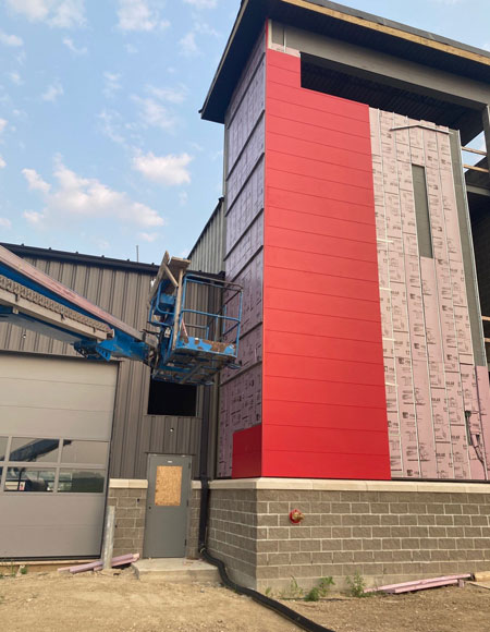 Construction site featuring a building with partially installed red wall panels, a lift for exterior work, and a gray brick foundation, illustrating commercial building envelope installation.