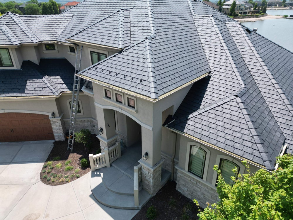 Residential roofing installation featuring DaVinci shingles on a private home in Waterloo, NE, with a ladder positioned against the house and a view of a nearby lake.